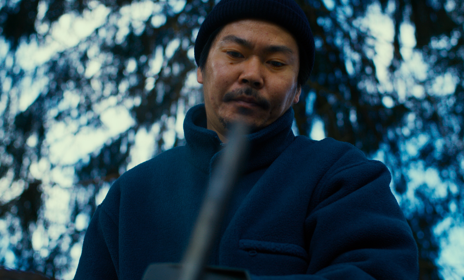 Takumi (Hitoshi Omika), a Japanese man in a watch cap and puffy coat, looks downward with an out-of-focus object in the foreground bisecting him in Ryûsuke Hamaguchi’s Evil Does Not Exist