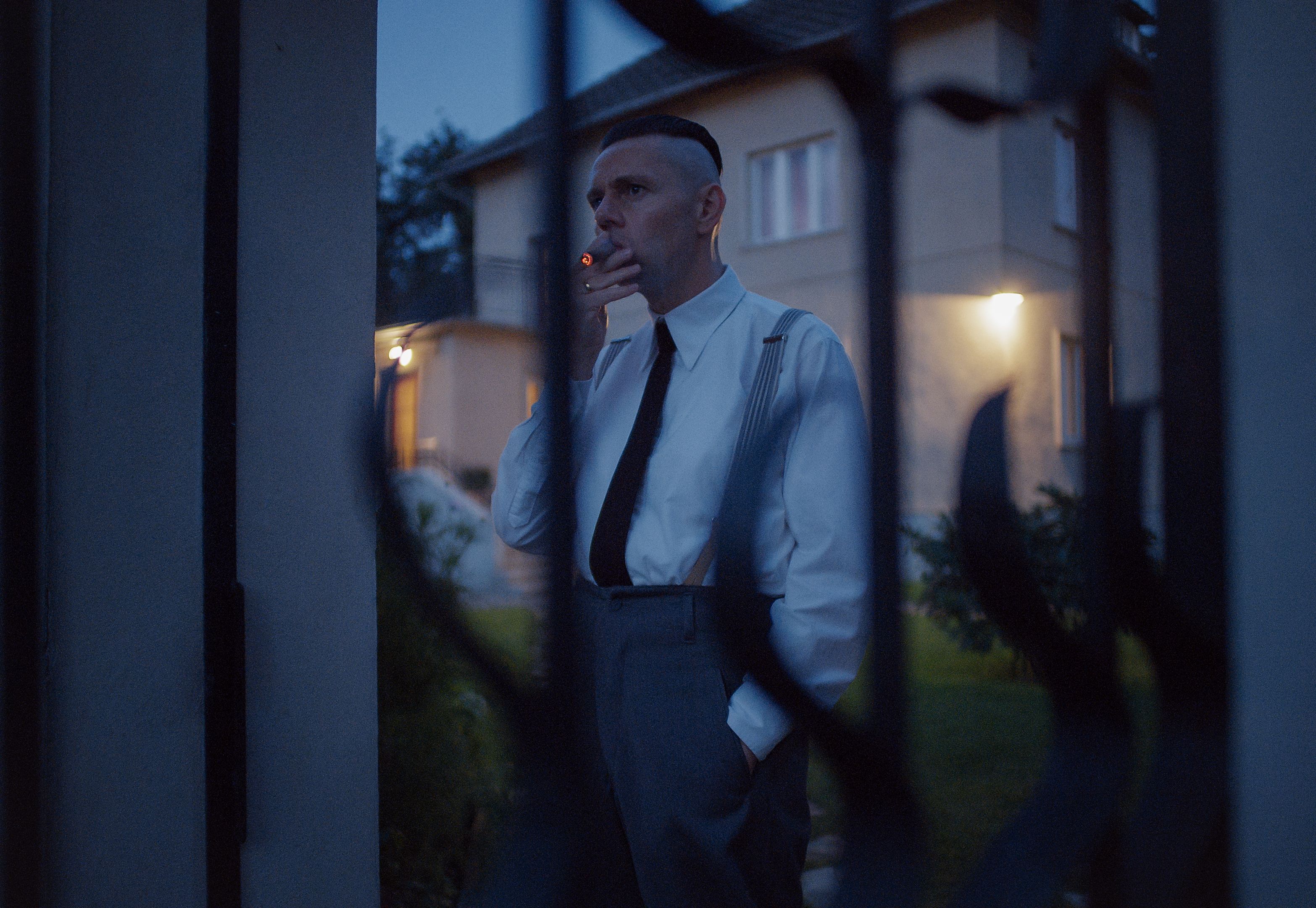 Christian Friedel as Rudolf Höss standing inside a gate and smoking a cigarette in The Zone of Interest