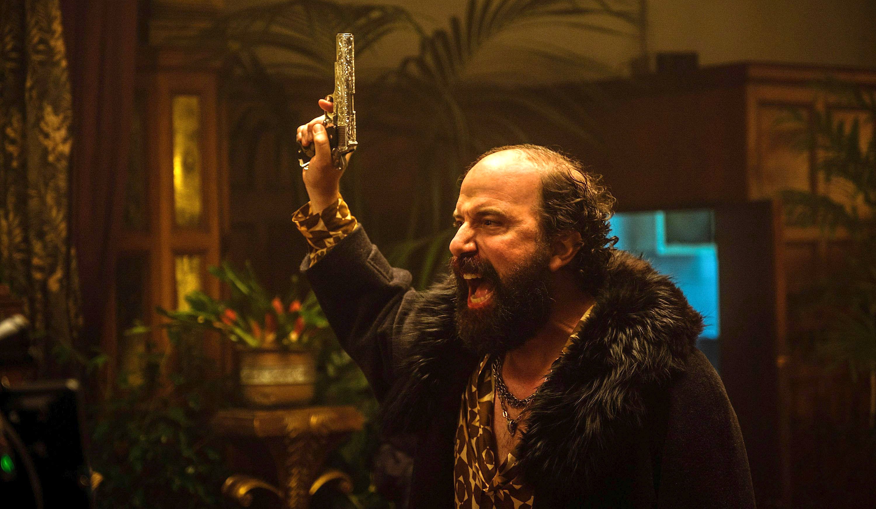 Gideon Van Der Koy (Brett Gelman), a balding, bearded man in a big, dark fur coat, yells and waves a gun in the air in Boy Kills World