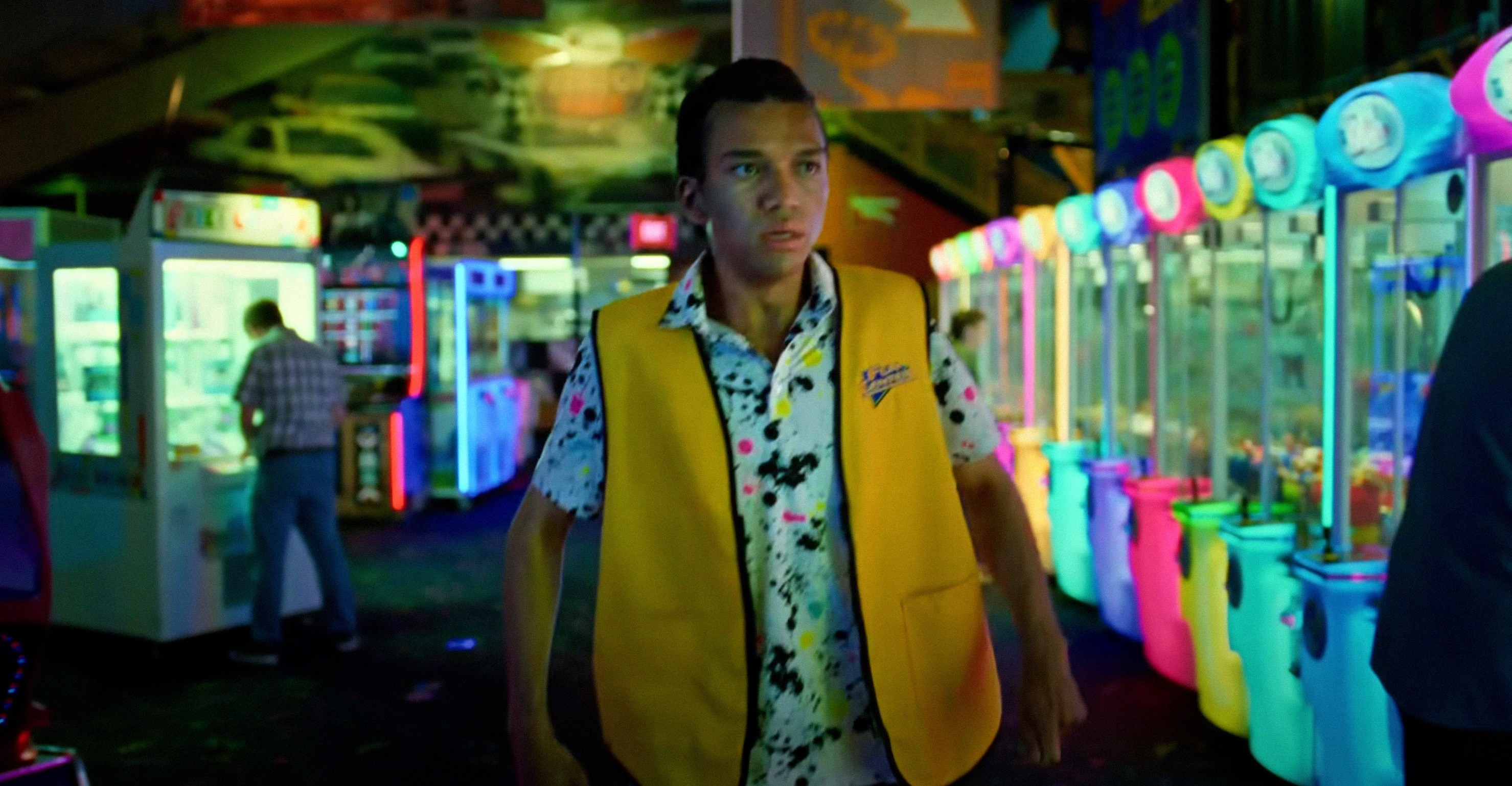 A middle-aged version of Owen (Justice Smith) in a button-up shirt and high-viz yellow vest walks through a dimly lit arcade with neon game machines all around him in Jane Schoenbrun’s I Saw the TV Glow