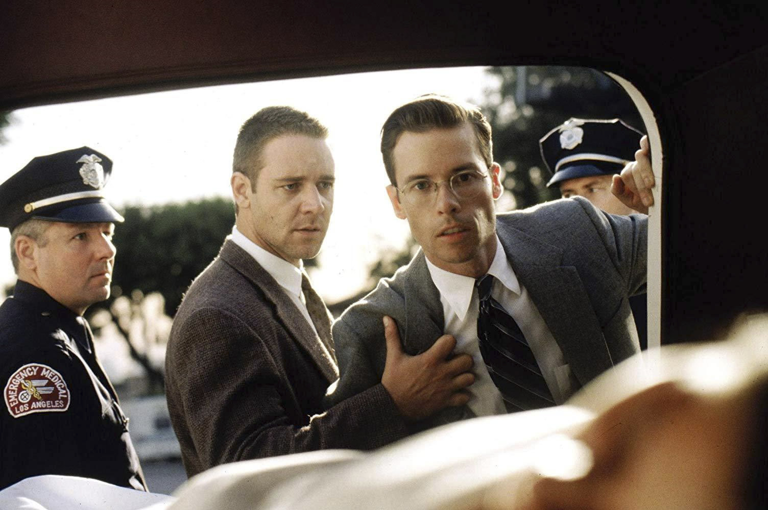 A man in a brown suit standing by a pair of police officer holding a man in a grey suit back as he stares into the back of a car with a body visible in the foreground.