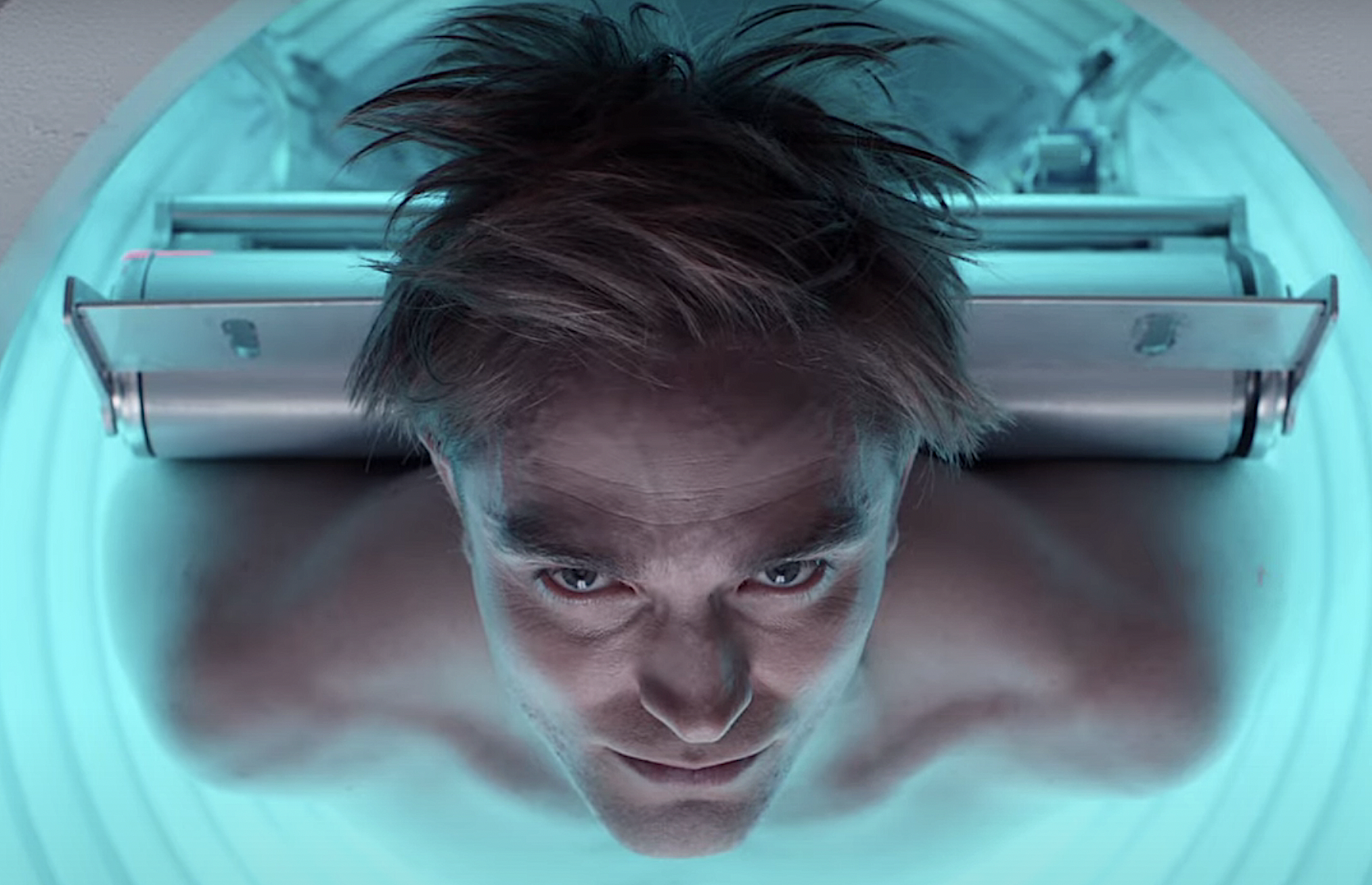 Robert Pattinson lies on his back, shirtless, in a glowing blue MRI-type machine, staring directly into the camera, in an early teaser trailer for Bong Joon-ho’s Mickey 17